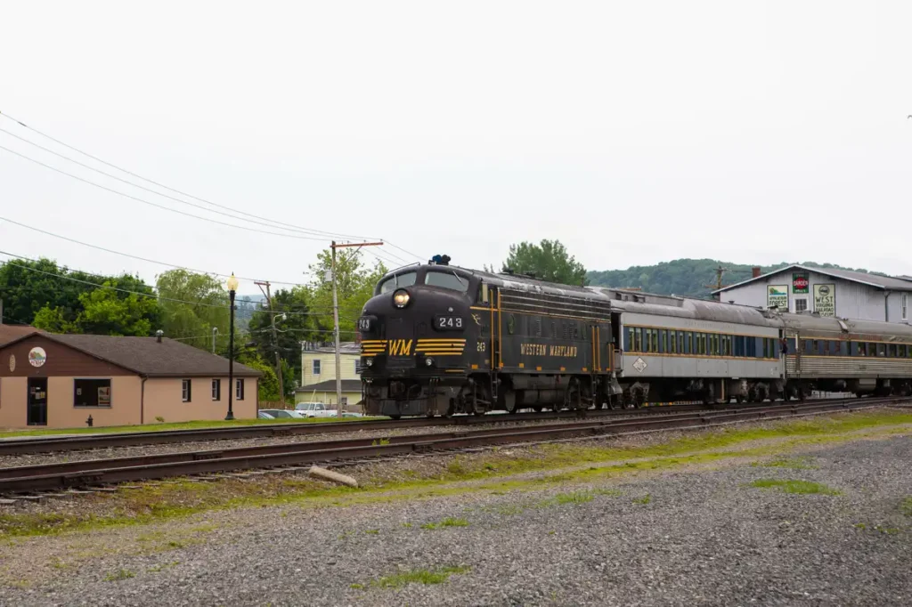 A vintage black and yellow Western Maryland scenic railroad train travels on tracks beside a building in downtown Elkins. The sky is overcast, and trees and hills are visible in the background.