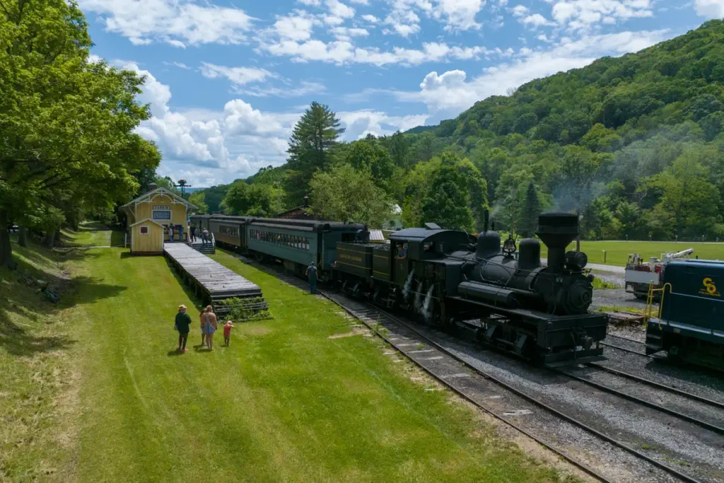 A Cass Scenic Railroad vintage steam train with dark carriages parked at a quaint station in Durbin under a bright sky. Three people walk along a grassy path, surrounded by lush trees and hills.