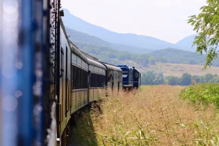A blue Potomac Eagle Scenic Railroad train winds through a grassy meadow against mountain and tree-lined horizon, evoking a sense of travel, adventure, and natural tranquility.