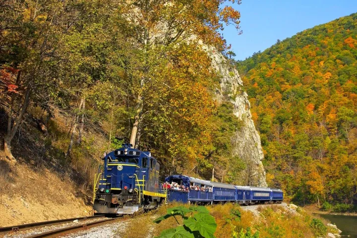 A vintage blue-and-yellow Potomac Eagle Scenic Railroad train travels through an autumnal forest canyon, surrounded by vibrant orange and green foliage on rocky hills, evoking adventure and nostalgia.