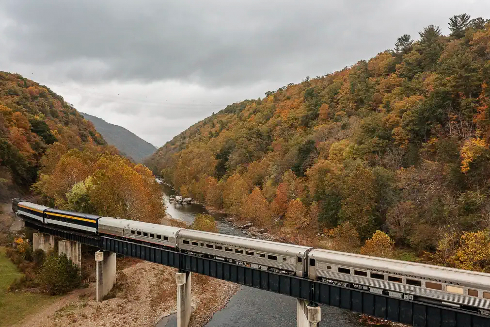 A Potomac Eagle Scenic Railroad train crosses a bridge over a river during autumn, surrounded by forested hills with colorful fall foliage under a cloudy sky.