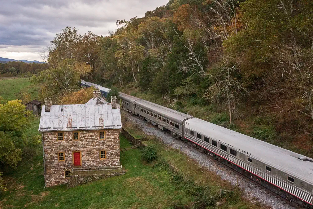 A vintage Potomac Eagle Scenic Railroad train passes by an old stone house with a red door, nestled in a lush, wooded landscape. The scene conveys a serene, nostalgic atmosphere.