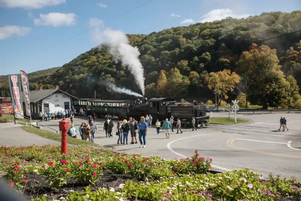 A Cass Scenic Railroad vintage steam train with billowing smoke is parked at a rural station. Visitors explore, surrounded by lush, autumn-colored hills and vibrant flowerbeds.