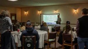 Elkins Board Meeting - Mon Forest Towns - Mon Forest Towns A man presents a slide titled Sustainable Tourism Indicators to a group of people seated at round tables in a dimly lit, wood-paneled conference room.