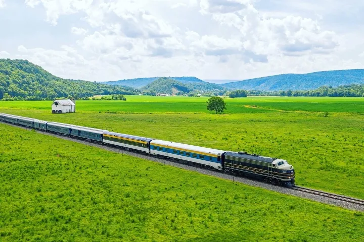 A scenic Potomac Eagle Railroad train travels through lush green fields with a mountainous backdrop under a partly cloudy sky. A small white barn is visible near the tracks.