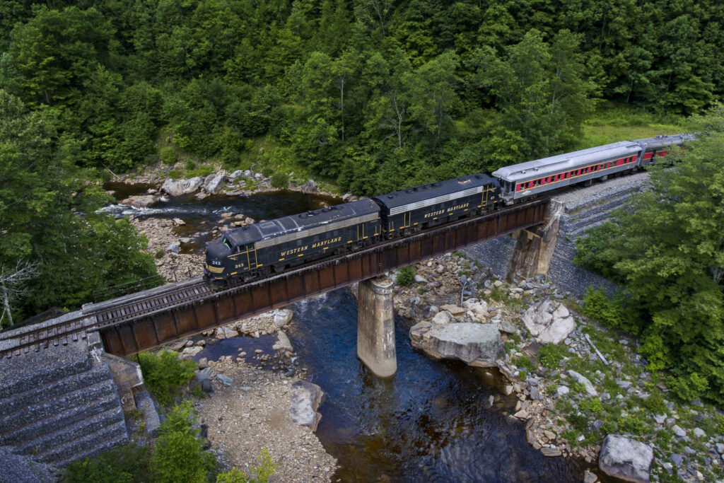 A vintage black and yellow scenic train crosses a rustic bridge over a rocky stream surrounded by lush green forest, conveying a serene and nostalgic atmosphere.