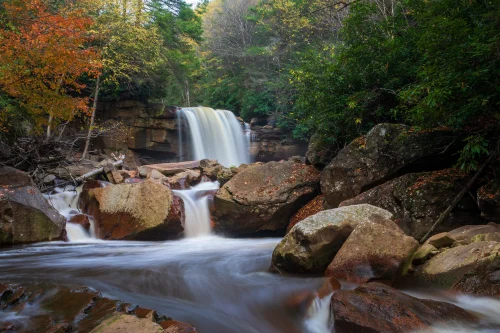 Waterfall flowing over a rocky cliff into a stream, surrounded by autumn trees with vibrant orange leaves.