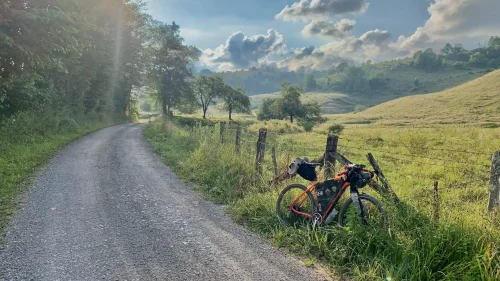 A gravel path winds through a lush, sunlit countryside with a bicycle leaning on a wooden fence. Rolling hills and fluffy clouds fill the serene landscape.